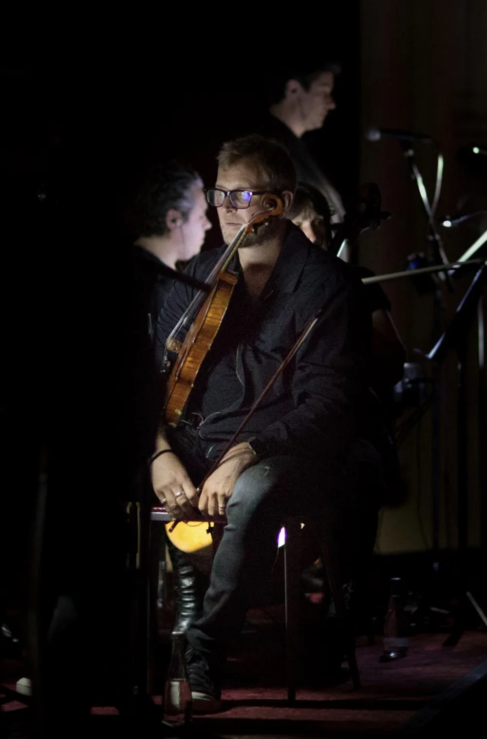 Bruce White seated on stage with his viola and bow, lit by a single spotlight among fellow musicians